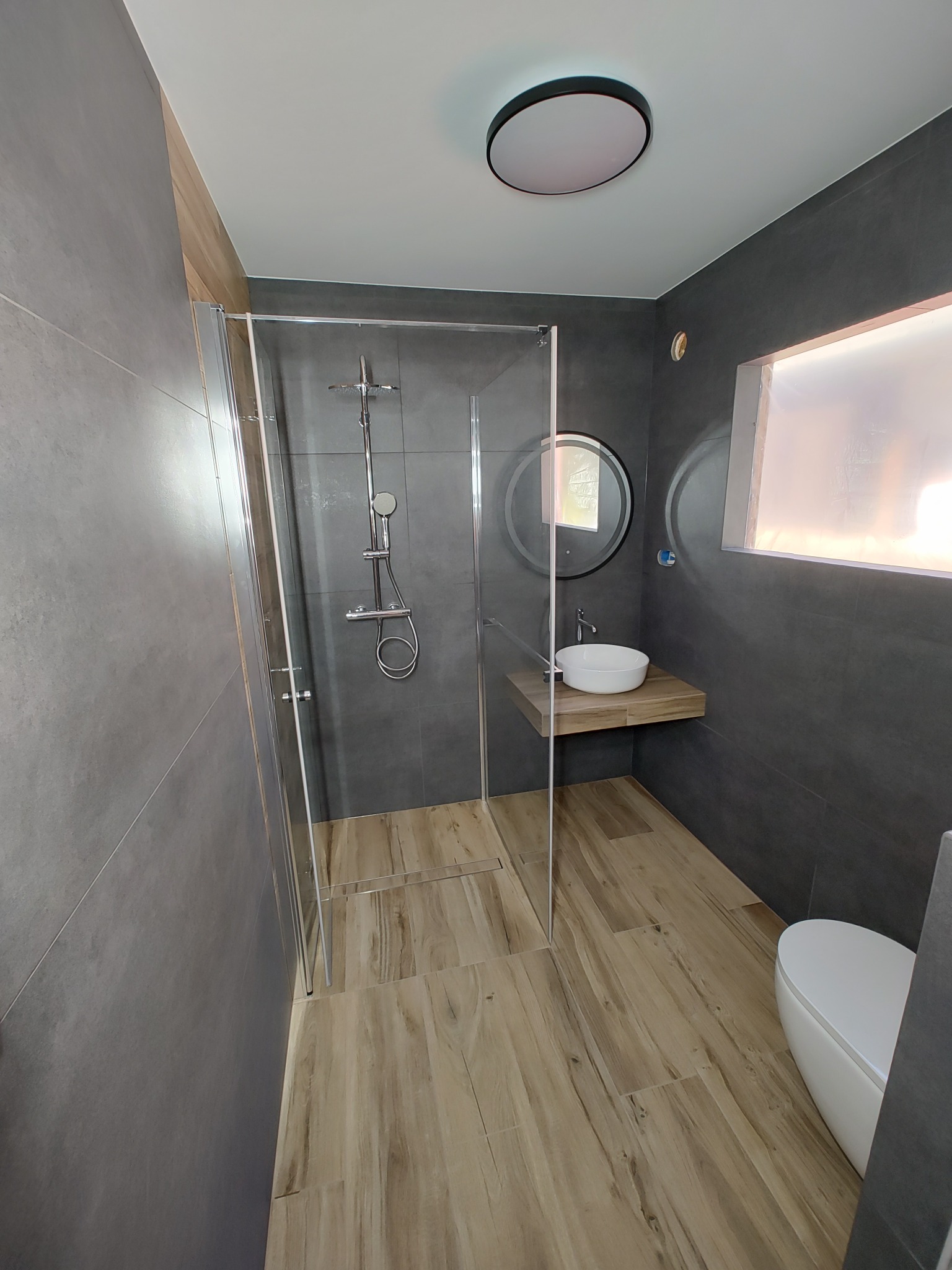 Nowoczesna łazienka z szarymi płytkami ściennymi, drewnopodobną podłogą, kabiną prysznicową ze szklanymi drzwiami, okrągłym lustrem z podświetleniem i białą umywalką nablatową na drewnianym blacie.