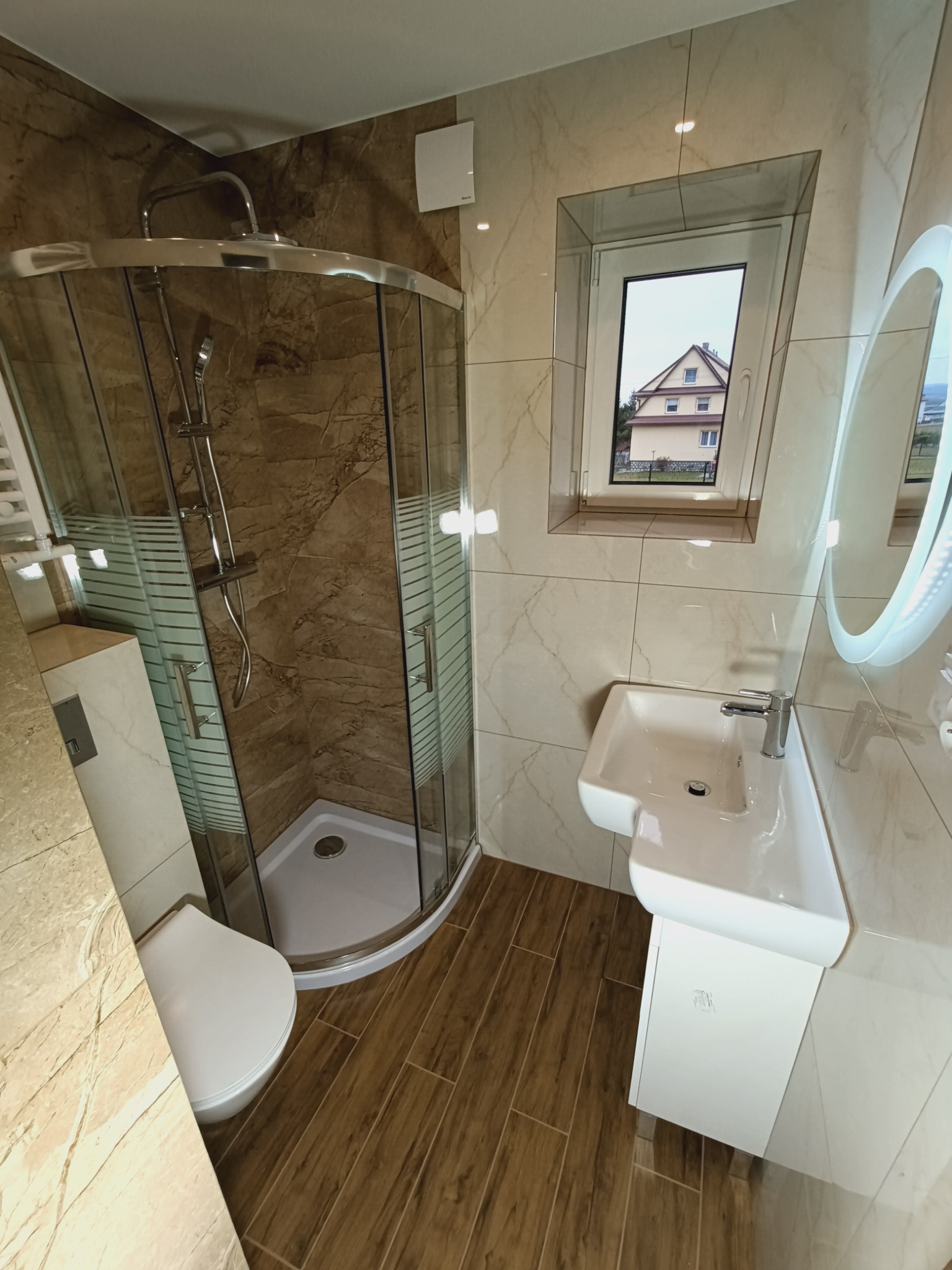 Nowoczesna łazienka z kabiną prysznicową narożną, umywalką na postumencie i podświetlanym lustrem, wykończona płytkami imitującymi marmur i drewno, z widokiem na dom przez okno.