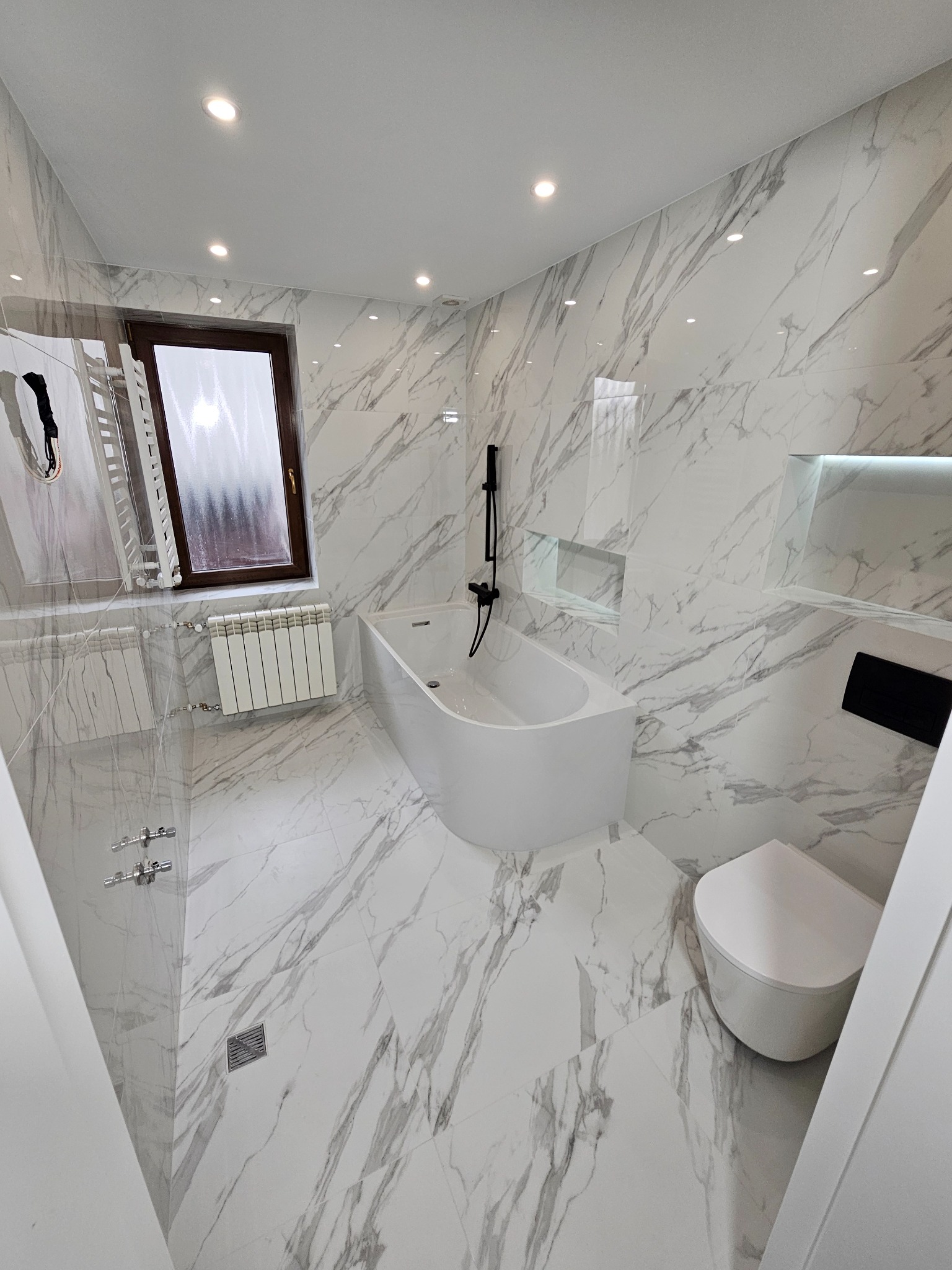 Nowoczesna łazienka wykończona białymi płytkami imitującymi marmur, z czarną armaturą i grzejnikiem drabinkowym, oświetlona punktowymi lampami sufitowymi.