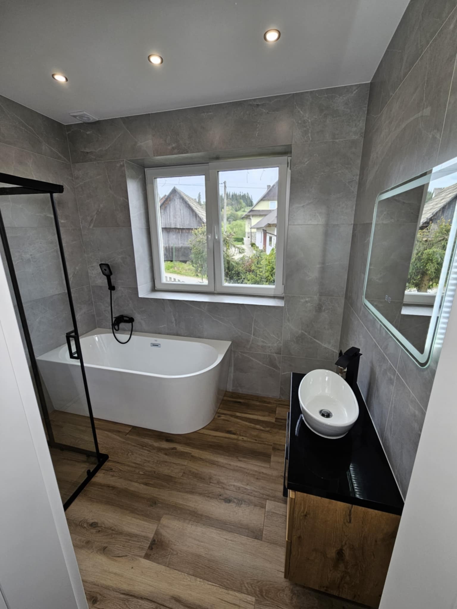 Nowoczesna łazienka z wanną wolnostojącą, szarą glazurą imitującą marmur i drewnopodobną podłogą. Okno z widokiem na wiejski krajobraz.