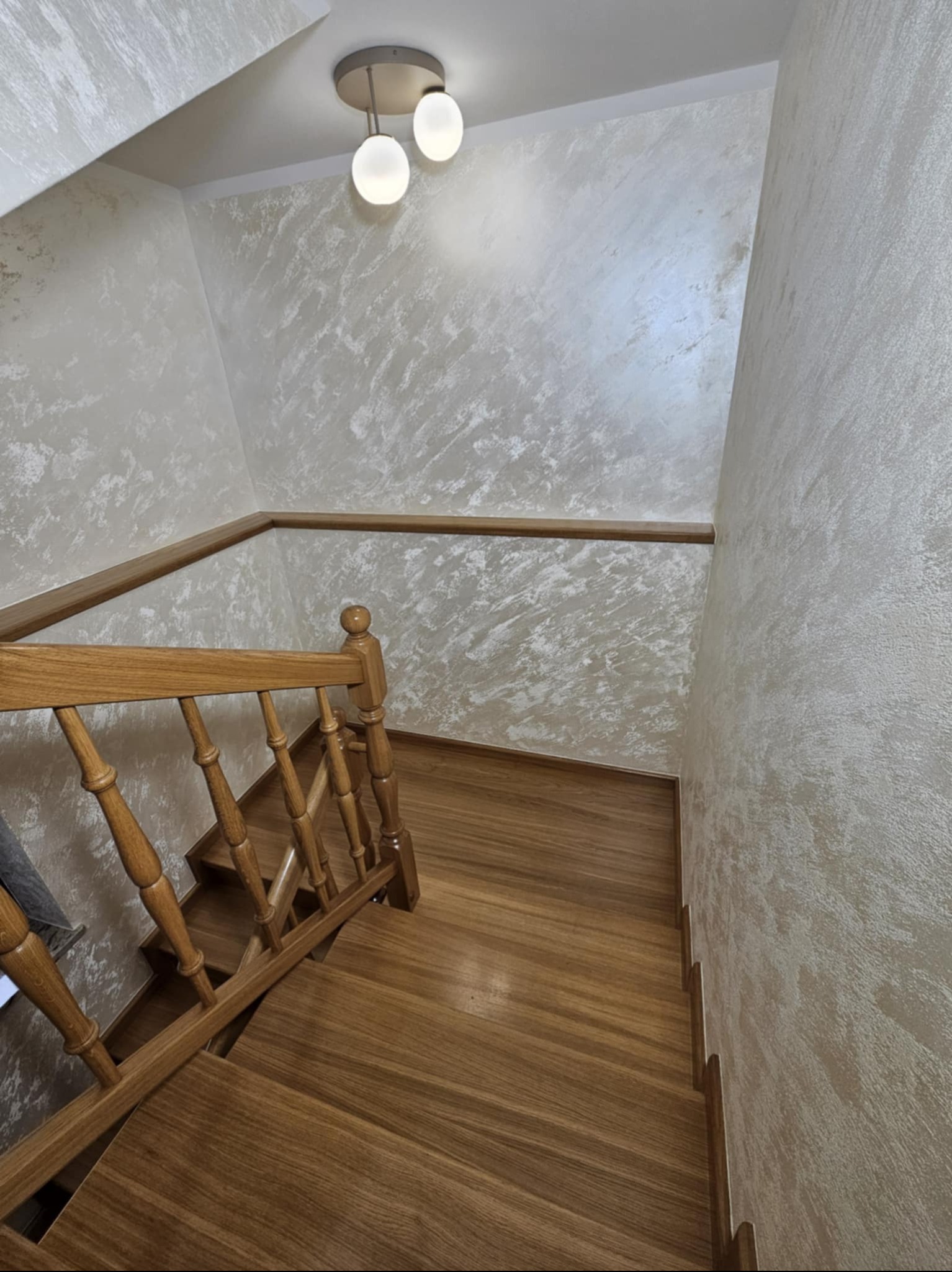 Klatka schodowa z drewnianymi schodami i balustradą. Ściany wykończone dekoracyjnym tynkiem w odcieniach beżu i bieli. Na suficie widoczna lampa z trzema kulistymi kloszami.