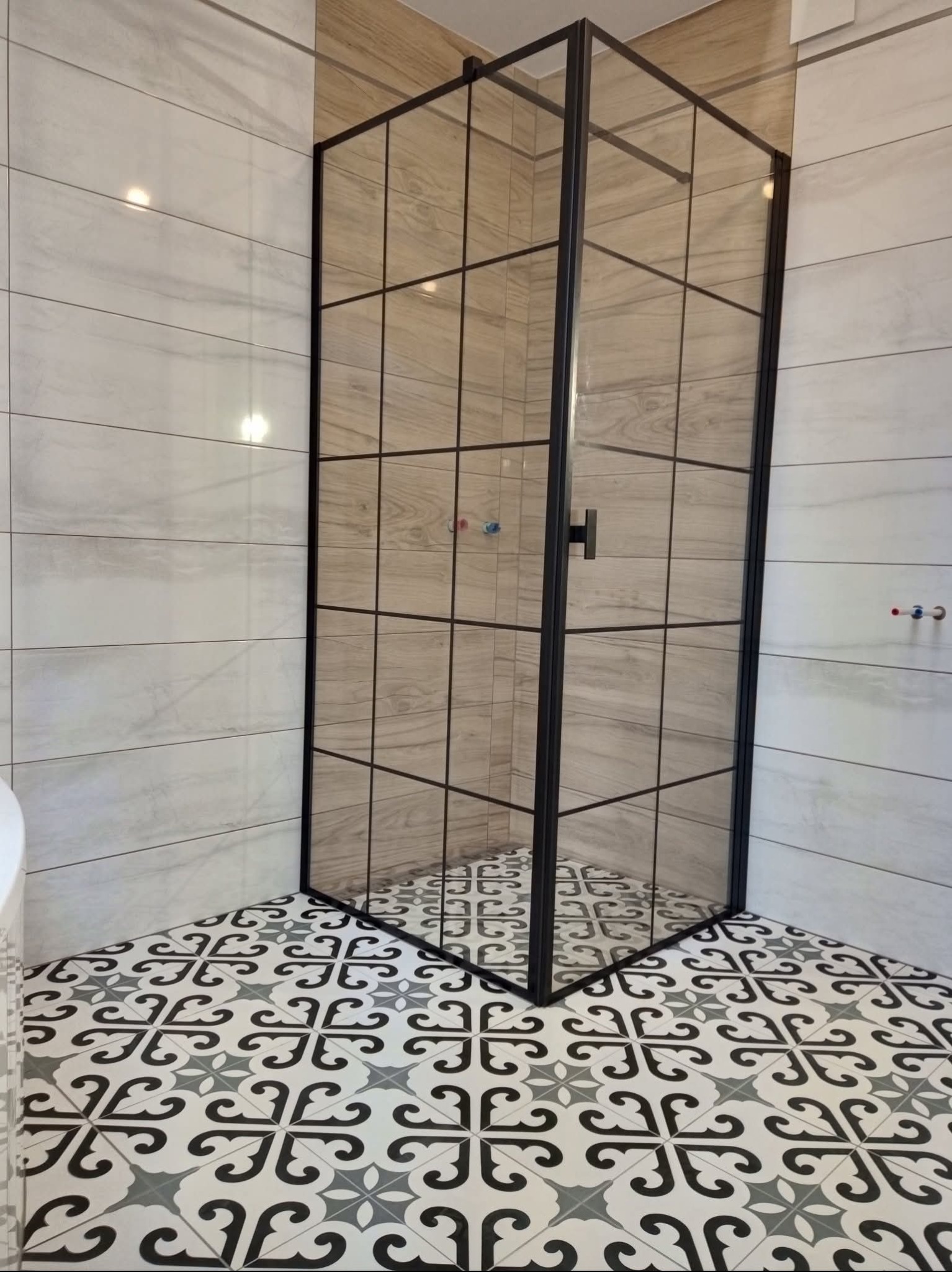 Nowoczesna kabina prysznicowa z czarnymi profilami i wzorzystą podłogą w wykończonej łazience. Ściany wyłożone płytkami imitującymi drewno i marmur.