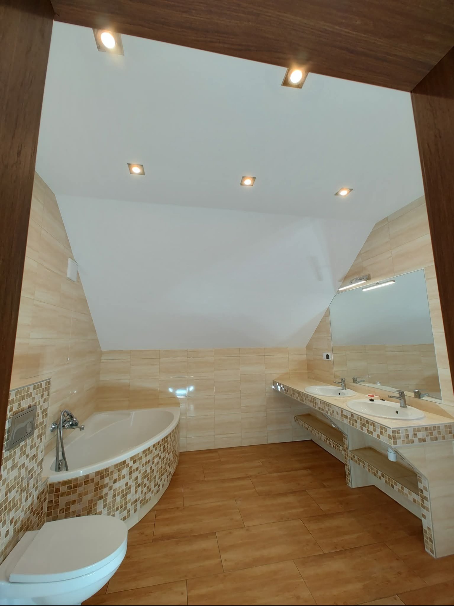 Jasna łazienka na poddaszu z mozaikową wanną, dwiema umywalkami i drewnopodobną podłogą. Widok zza drewnianej framugi drzwi.