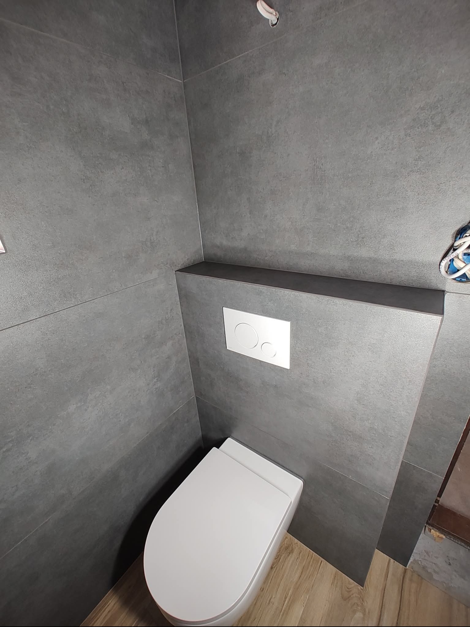 Nowoczesna toaleta podwieszana z szarą, matową okładziną ścienną i drewnopodobną podłogą. Minimalistyczny design, widoczny przycisk spłukujący.