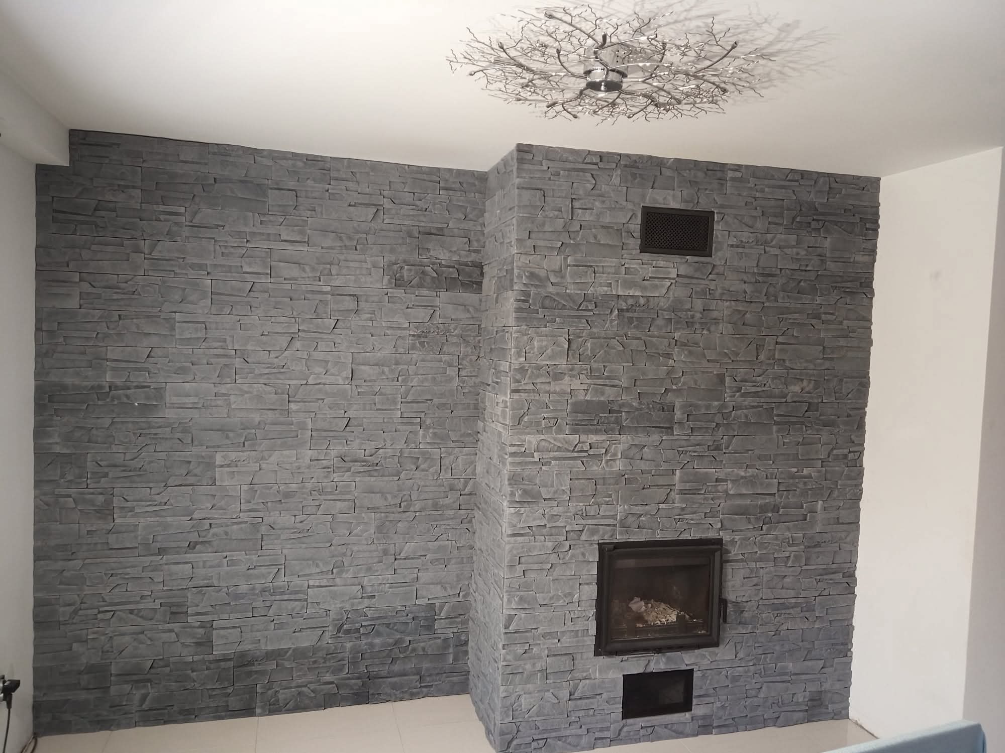 Ściana z szarej, dekoracyjnej cegły imitującej kamień, z wbudowanym kominkiem i kratką wentylacyjną. Na suficie ozdobna lampa w kształcie gałęzi.