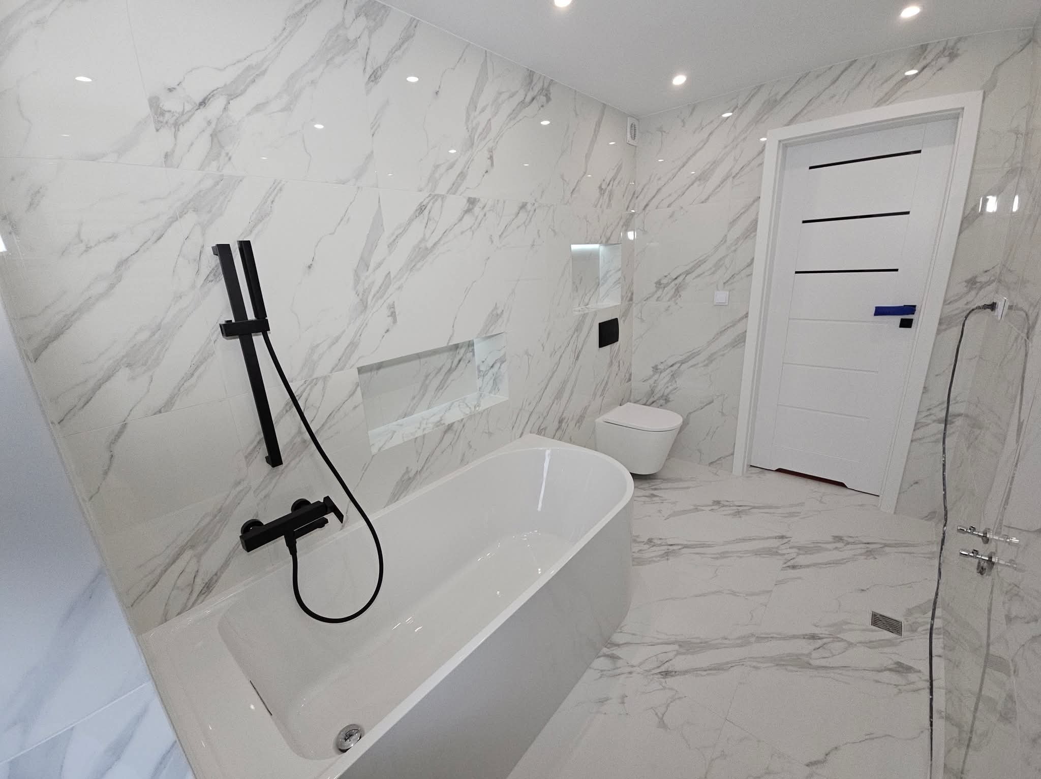 Nowoczesna łazienka z białymi płytkami imitującymi marmur, czarną armaturą i minimalistyczną toaletą podwieszaną. Widoczna wanna i drzwi z czarnymi akcentami.