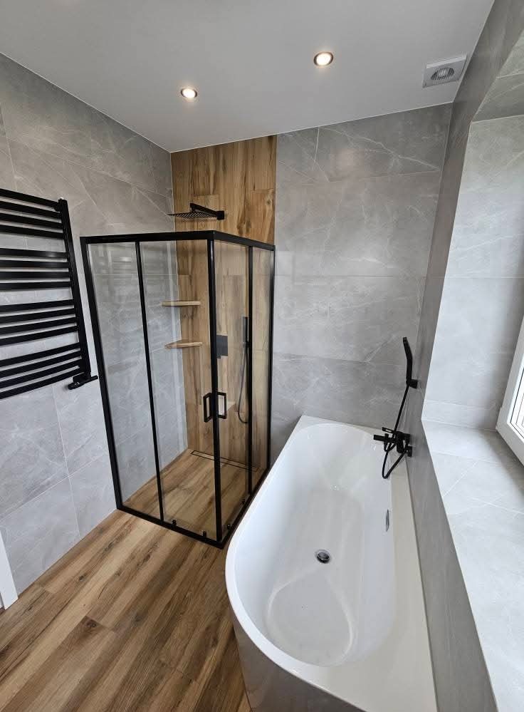 Nowoczesna łazienka z kabiną prysznicową typu walk-in z czarnymi ramami, białą wanną i grzejnikiem drabinkowym na tle szarych płytek imitujących marmur.
