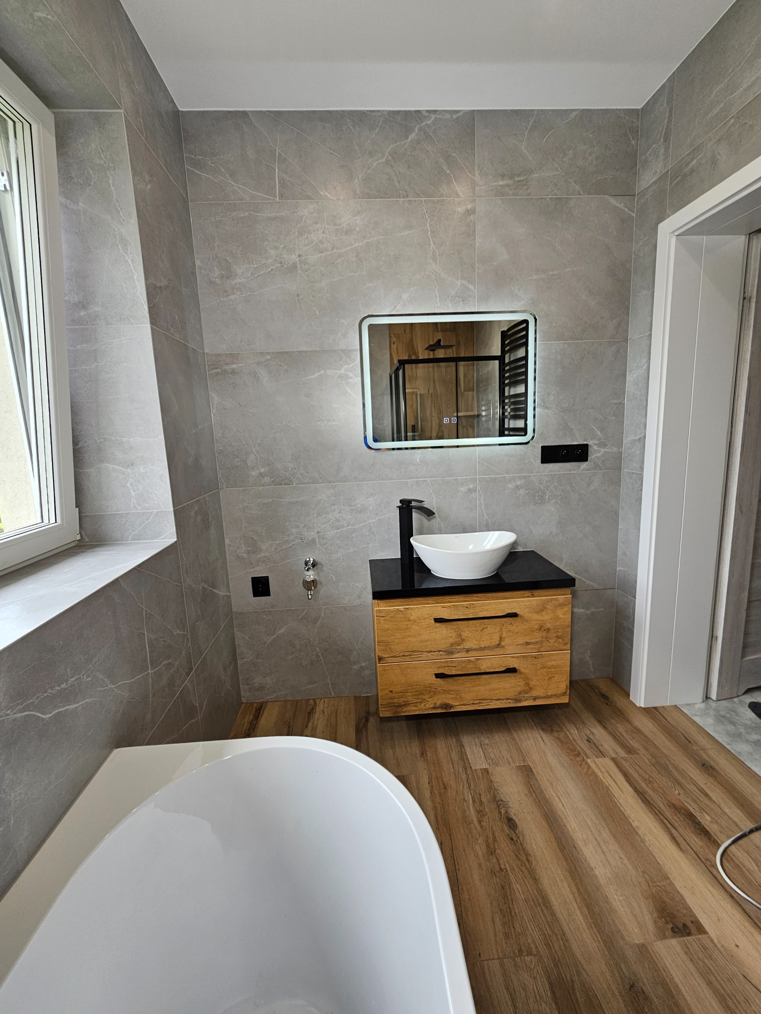 Nowoczesna łazienka z szarymi płytkami imitującymi marmur, drewnianą szafką pod umywalką i podświetlanym lustrem. Widoczna wanna i drewnopodobna podłoga.