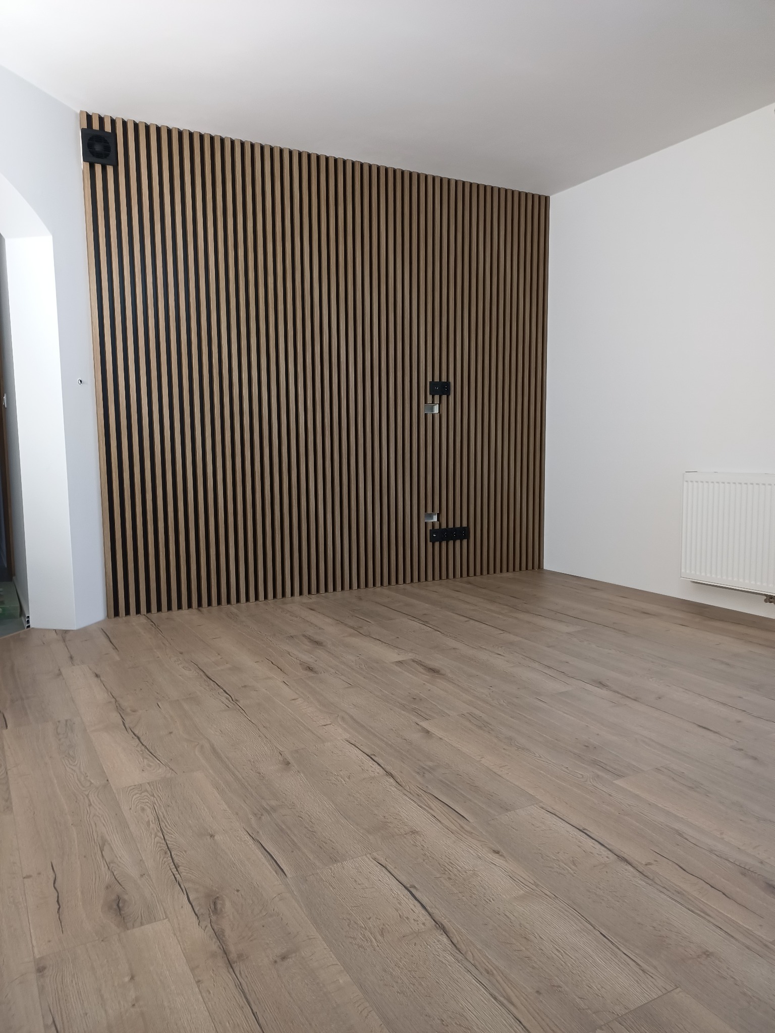 Nowoczesne wykończenie wnętrza: drewniane lamele na ścianie z czarnymi akcentami, jasna podłoga z paneli, białe ściany i grzejnik. Minimalistyczny design.