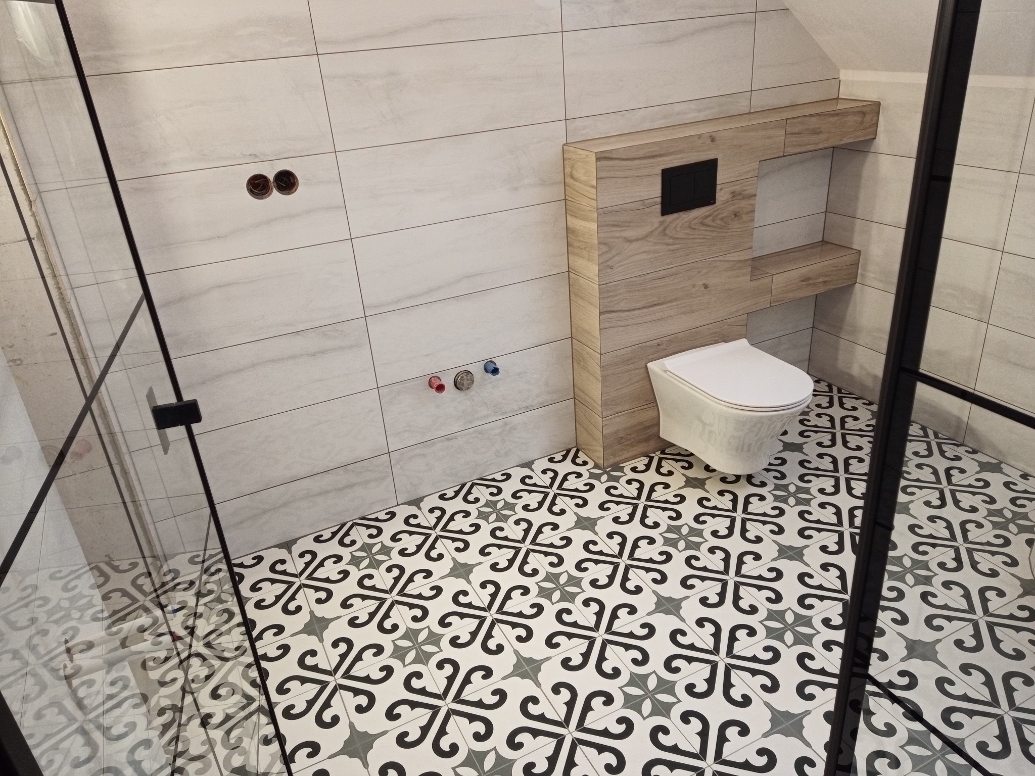 Wykończona łazienka w odcieniach bieli i szarości z wzorzystą podłogą. Widoczna toaleta z zabudową z drewnopodobnych paneli i fragment kabiny prysznicowej z czarnymi ramami.