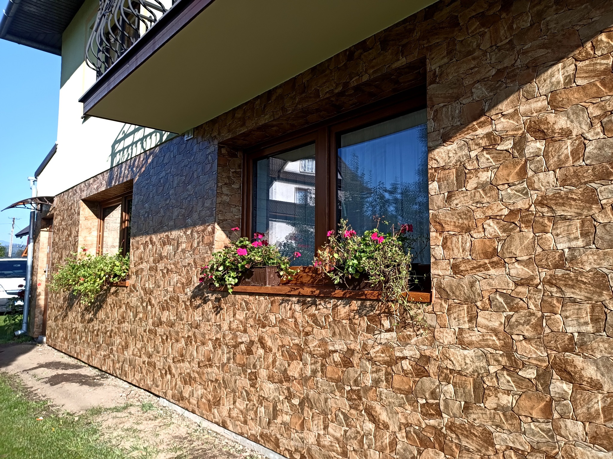 Elewacja budynku wykończona kamieniem dekoracyjnym w odcieniach brązu i beżu. Okna z drewnianą ramą, ozdobione donicami z kwiatami. Widok z boku, w słoneczny dzień.