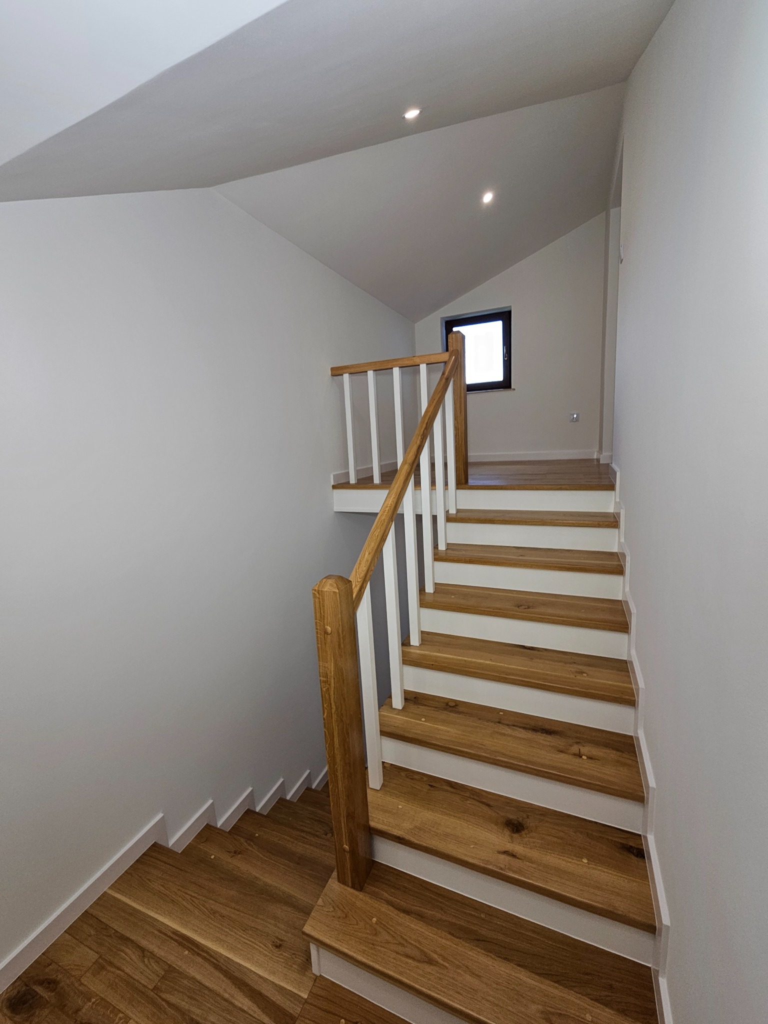 Drewniane schody z białymi podstopnicami i balustradą w jasnym wnętrzu. Widoczne oświetlenie punktowe i okno. Minimalistyczny design.