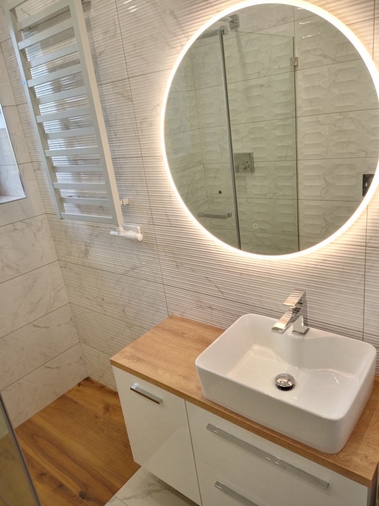 Nowoczesna łazienka z umywalką nablatową, podświetlanym lustrem i grzejnikiem drabinkowym na tle płytek imitujących marmur. Widoczna kabina prysznicowa.