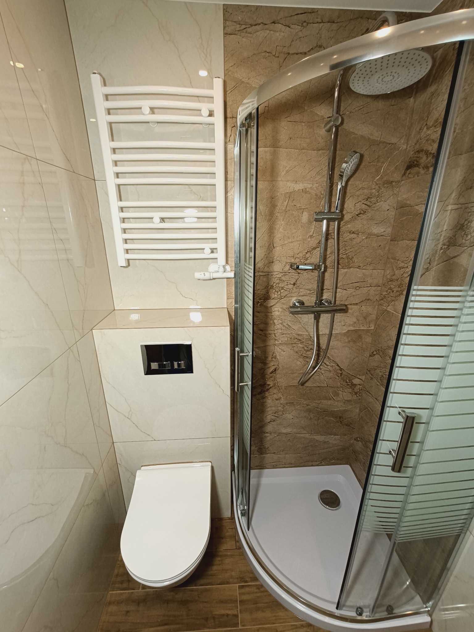 Nowoczesna łazienka z kabiną prysznicową, podwieszaną toaletą i grzejnikiem drabinkowym. Beżowe płytki imitujące marmur na ścianach i drewnopodobne na podłodze.