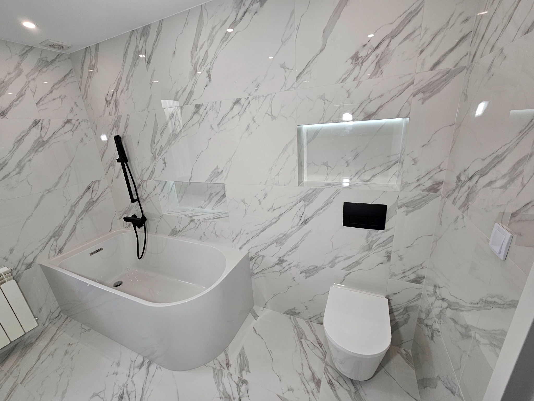 Elegancka łazienka w Podwilku z wanną, toaletą i płytkami imitującymi marmur. Czarna armatura i oświetlenie wnęki dodają nowoczesnego charakteru.