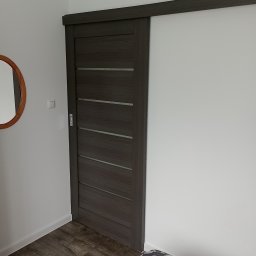 Dawid Ćwir - Zamontowane przesuwne drzwi w odcieniach szarości z poziomymi wstawkami, widoczne w pokoju z drewnopodobną podłogą i okrągłym lustrem na ścianie.
