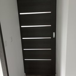 Dawid Ćwir - Ciemne, przesuwne drzwi z poziomymi, jasnymi wstawkami, zamontowane w białej ścianie, widoczne z perspektywy podłogi.