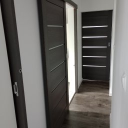 Dawid Ćwir - Ciemne, przesuwne drzwi z poziomymi przeszkleniami, widoczne w korytarzu z panelami podłogowymi, z drugimi drzwiami w tle.