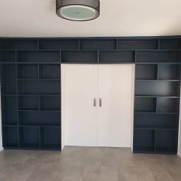 Dawid Ćwir - Wnęka z zabudowanymi, ciemnoniebieskimi regałami po bokach białych, dwuskrzydłowych drzwi, szara podłoga, widoczna lampa sufitowa.