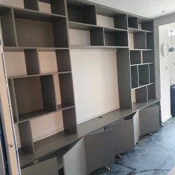 Dawid Ćwir - Szara zabudowa meblowa z otwartymi i zamkniętymi półkami, fronty szafek częściowo otwarte, widoczne podpórki montażowe na podłodze.