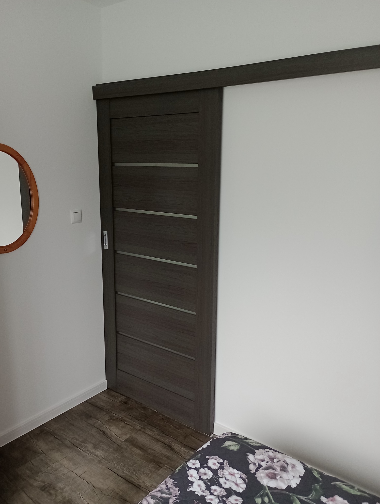 Zamontowane przesuwne drzwi w odcieniach szarości z poziomymi wstawkami, widoczne w pokoju z drewnopodobną podłogą i okrągłym lustrem na ścianie.