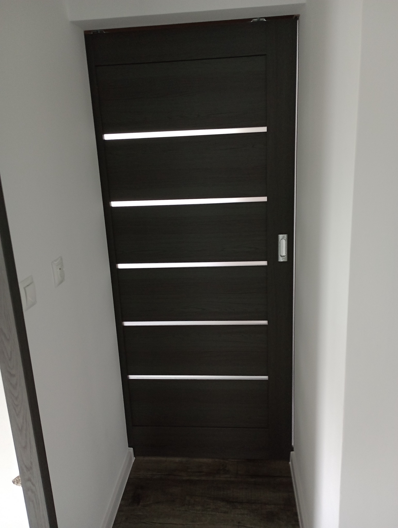 Ciemne, przesuwne drzwi z poziomymi, jasnymi wstawkami, zamontowane w białej ścianie, widoczne z perspektywy podłogi.