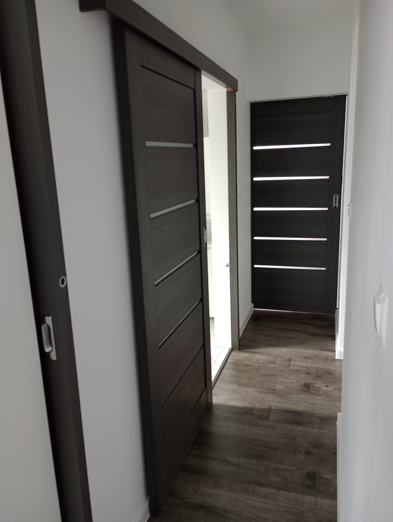 Ciemne, przesuwne drzwi z poziomymi przeszkleniami, widoczne w korytarzu z panelami podłogowymi, z drugimi drzwiami w tle.