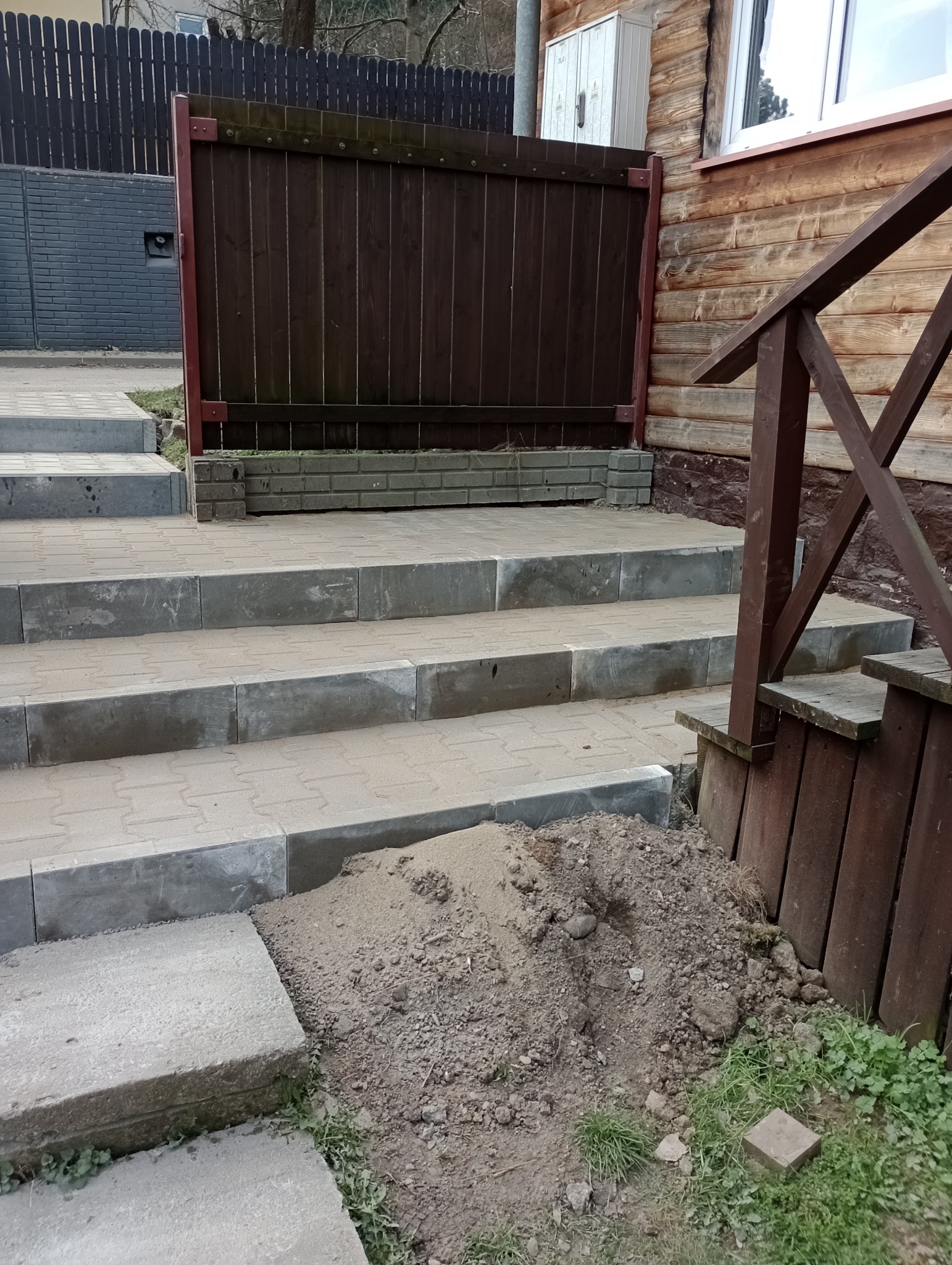 Schody z kostki brukowej prowadzące do drewnianego domu, z widoczną stertą ziemi i fragmentem drewnianego ogrodzenia.
