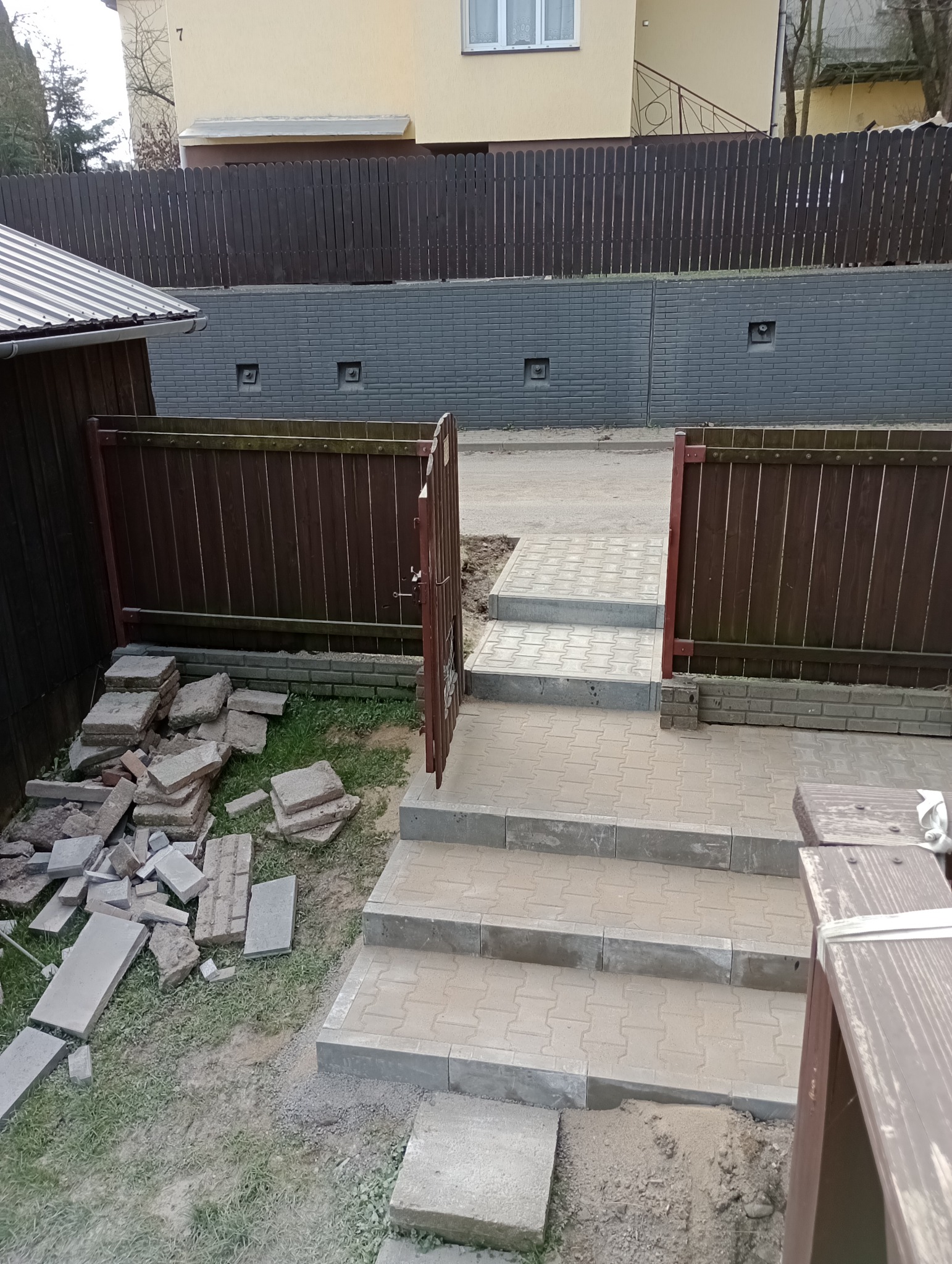 Schody zewnętrzne z kostki brukowej prowadzące do furtki w brązowym płocie, obok stosy nieułożonej kostki.