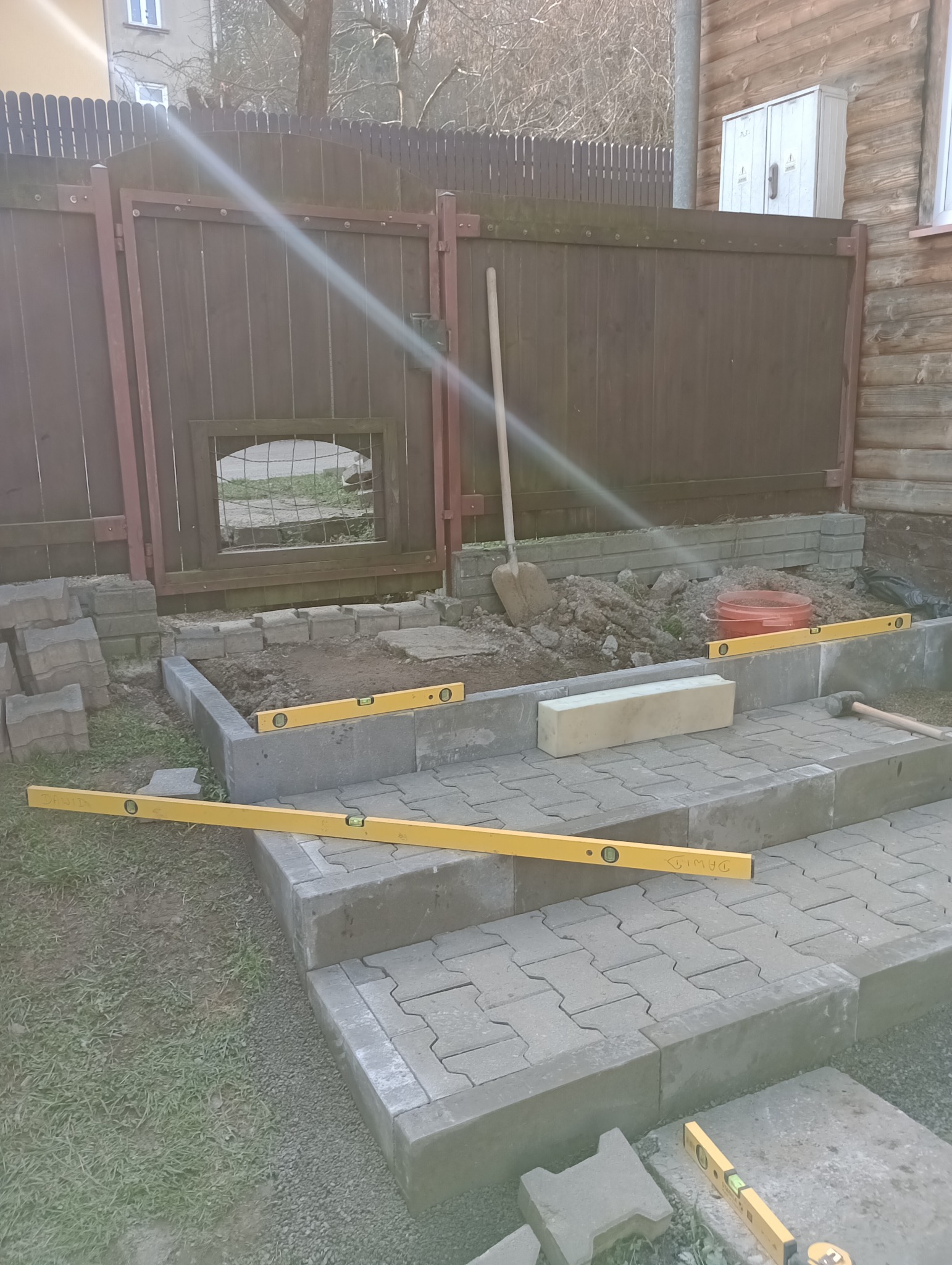 Budowa schodów zewnętrznych z kostki brukowej, poziomice na stopniach, widoczna łopata i fragment ogrodzenia.