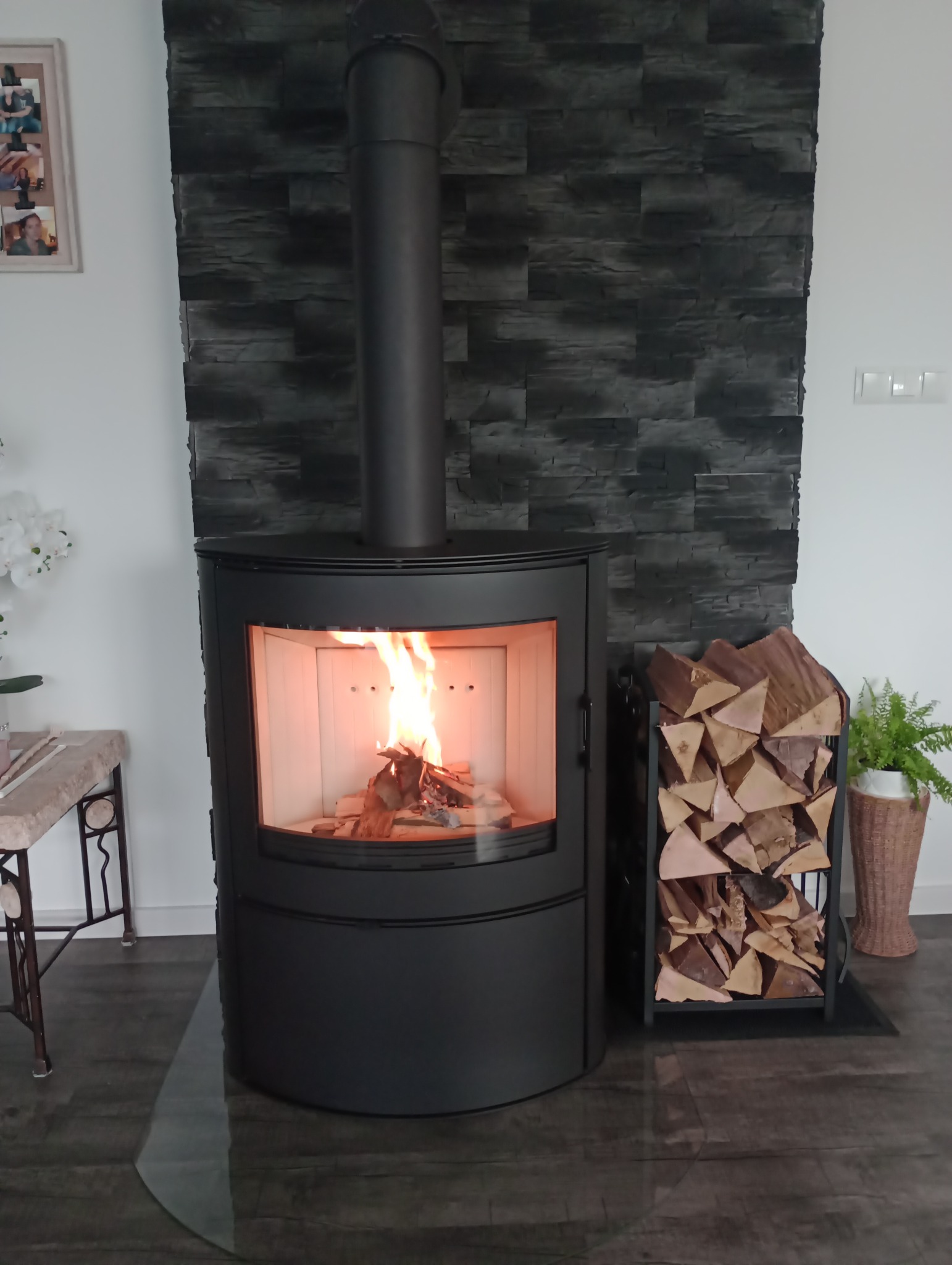 Nowoczesny, czarny piec wolnostojący z palącym się drewnem w jasnym wnętrzu, obok stojak na drewno i roślina doniczkowa, ściana wykończona okładziną imitującą kamień.