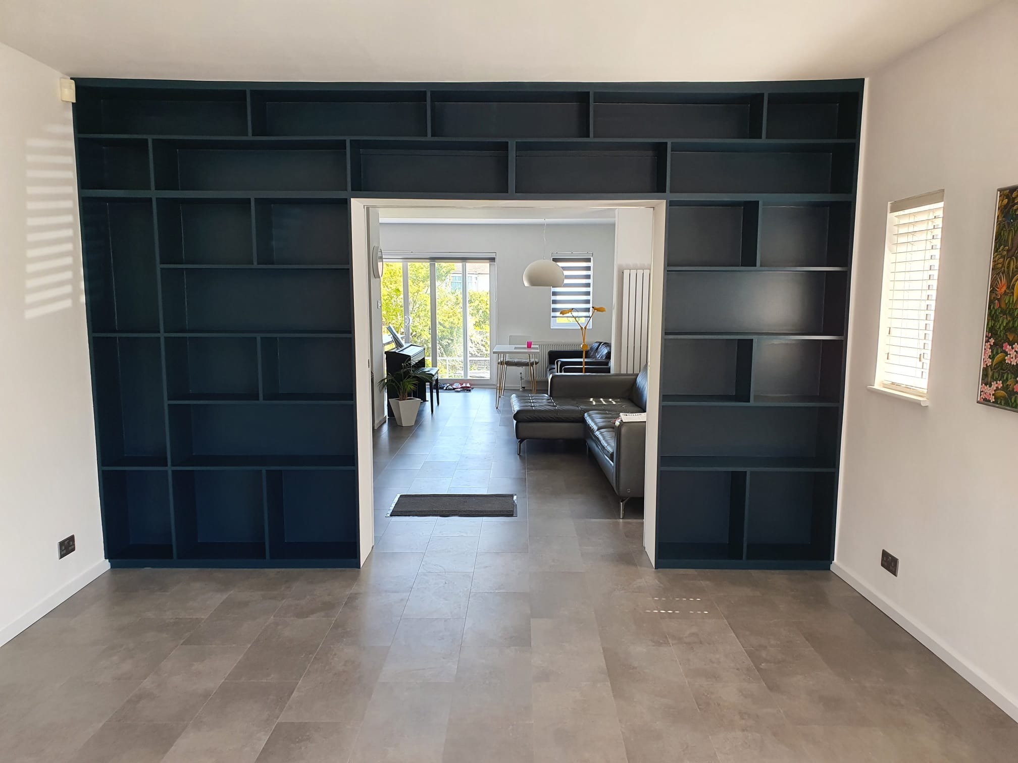 Przestronny salon z szarą podłogą i wbudowanym, ciemnogranatowym regałem na całej ścianie, tworzącym przejście do drugiego pokoju z widocznym pianinem i skórzaną sofą.