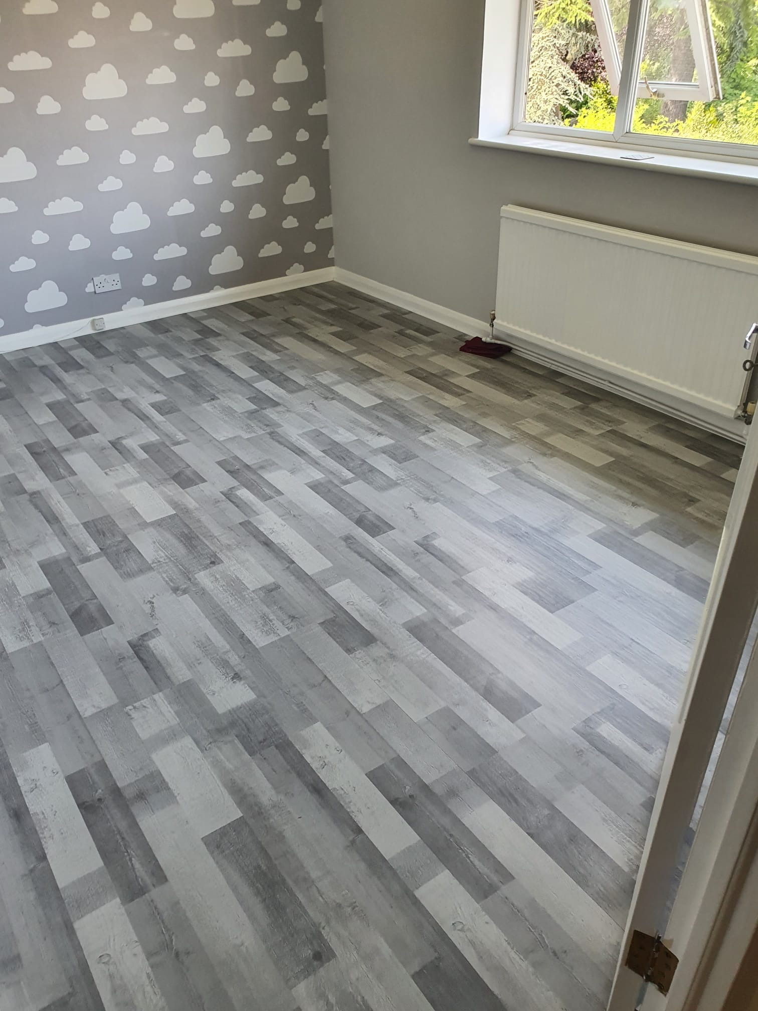 Pokój po remoncie z szarą podłogą imitującą drewno ułożoną w jodełkę, ściana z motywem chmur, biały grzejnik pod oknem.