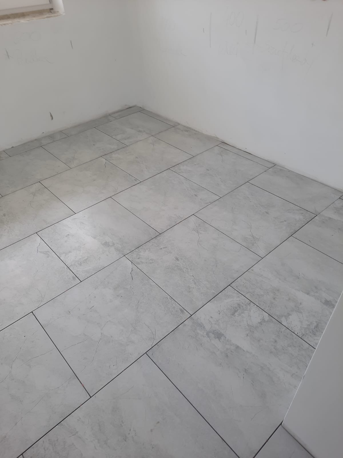 Świeżo ułożona, jasnoszara podłoga z płytek imitujących marmur w rogu pomieszczenia z surowymi, białymi ścianami z widocznymi notatkami ołówkiem.