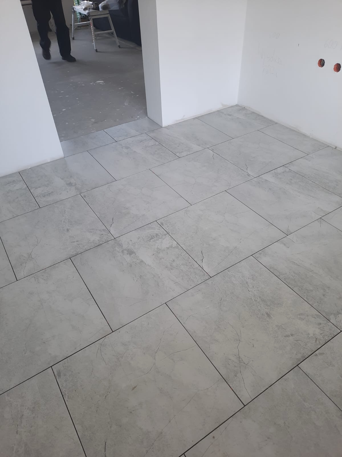 Ułożone szare płytki podłogowe imitujące kamień w nowym pomieszczeniu, widoczne ślady po pracach wykończeniowych na ścianach, w tle fragment nogi osoby i sprzęt.
