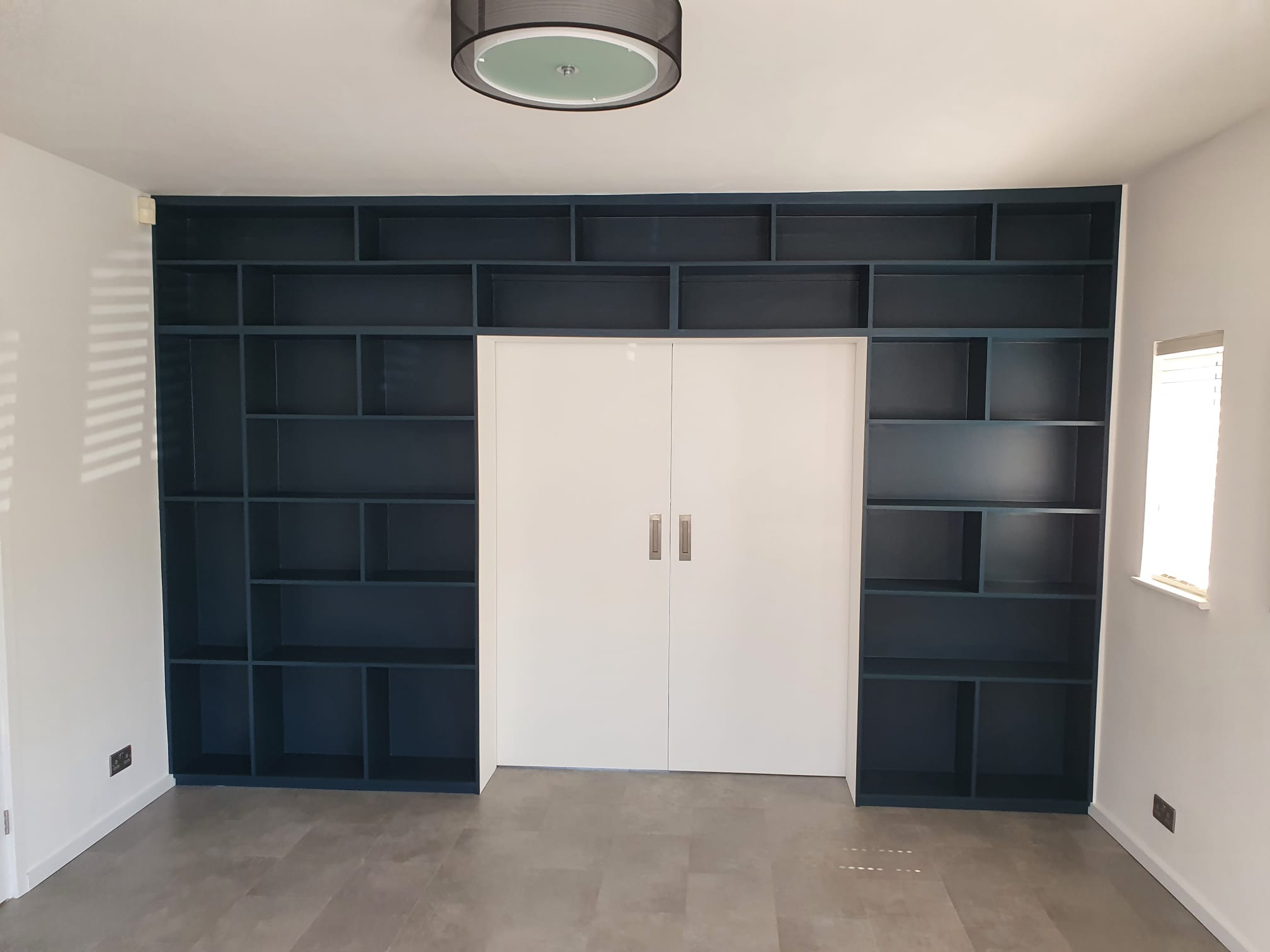 Wnęka z zabudowanymi, ciemnoniebieskimi regałami po bokach białych, dwuskrzydłowych drzwi, szara podłoga, widoczna lampa sufitowa.