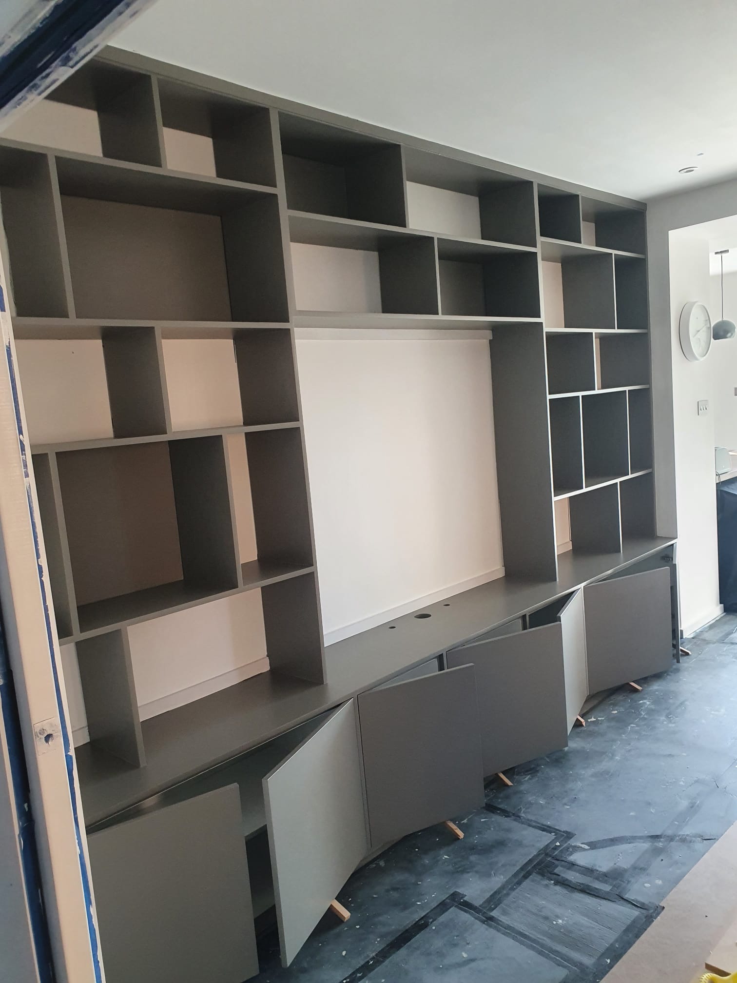 Szara zabudowa meblowa z otwartymi i zamkniętymi półkami, fronty szafek częściowo otwarte, widoczne podpórki montażowe na podłodze.