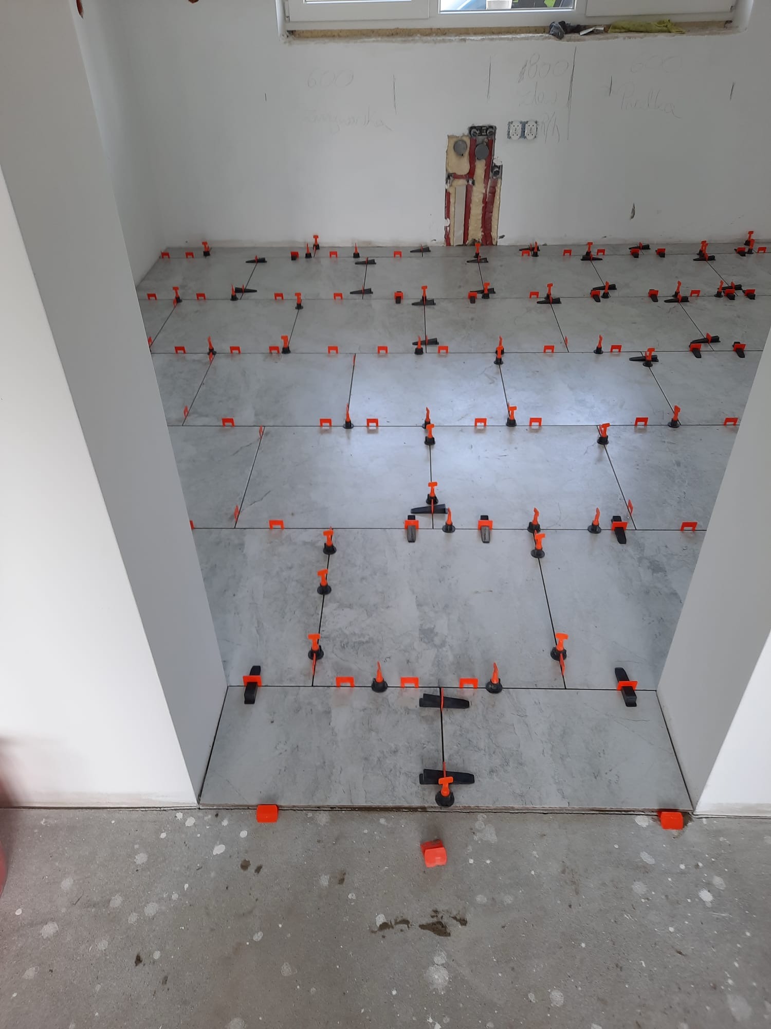 Układanie szarych płytek podłogowych z użyciem pomarańczowych klipsów poziomujących, widoczne niedokończone ściany z instalacją elektryczną i zapiskami.