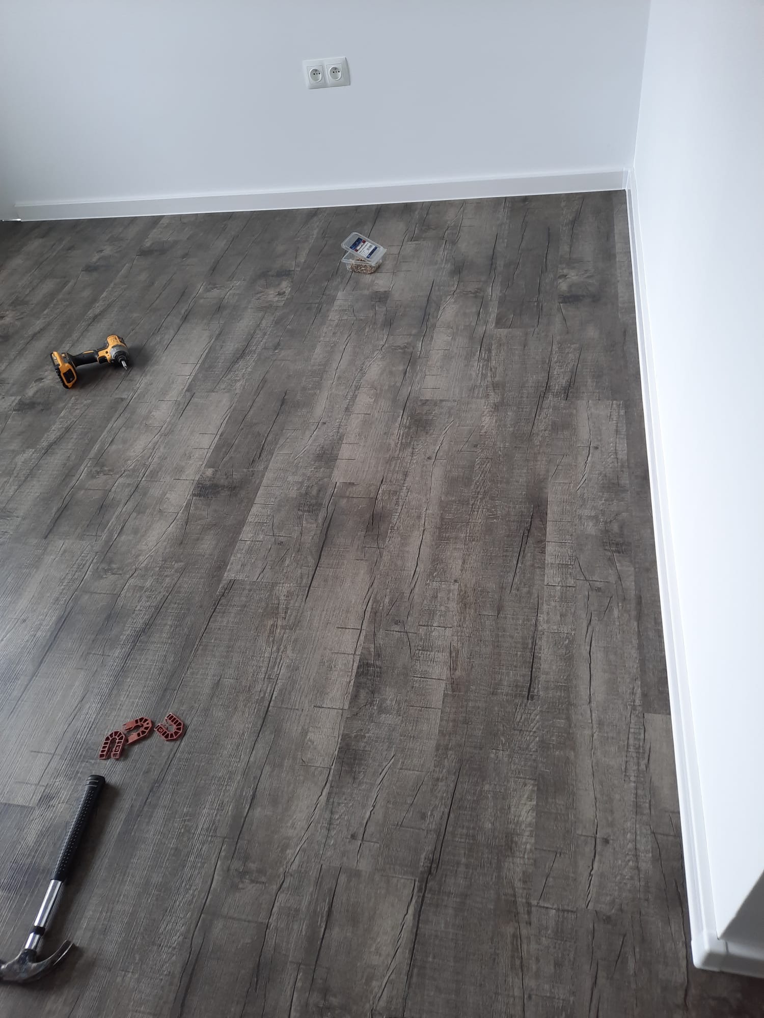 Szare panele podłogowe imitujące drewno, częściowo ułożone, widoczne narzędzia: młotek, wkrętarka, dystanse do paneli i pudełko z kołkami, białe ściany z listwą przypodłogową i gniazdkami.