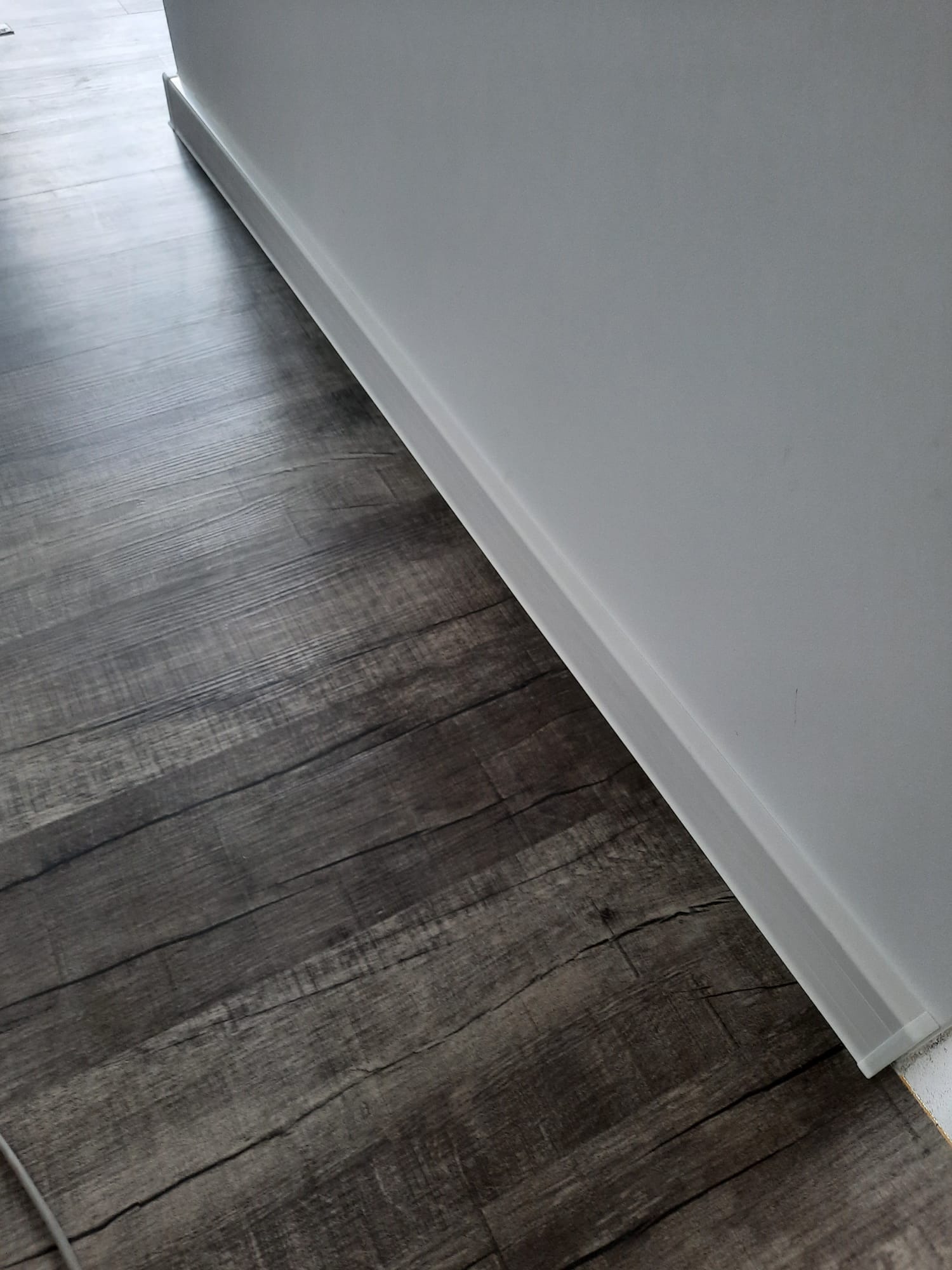 Szare panele podłogowe imitujące drewno, stykające się z białą ścianą i listwą przypodłogową, widoczne z niskiego kąta.
