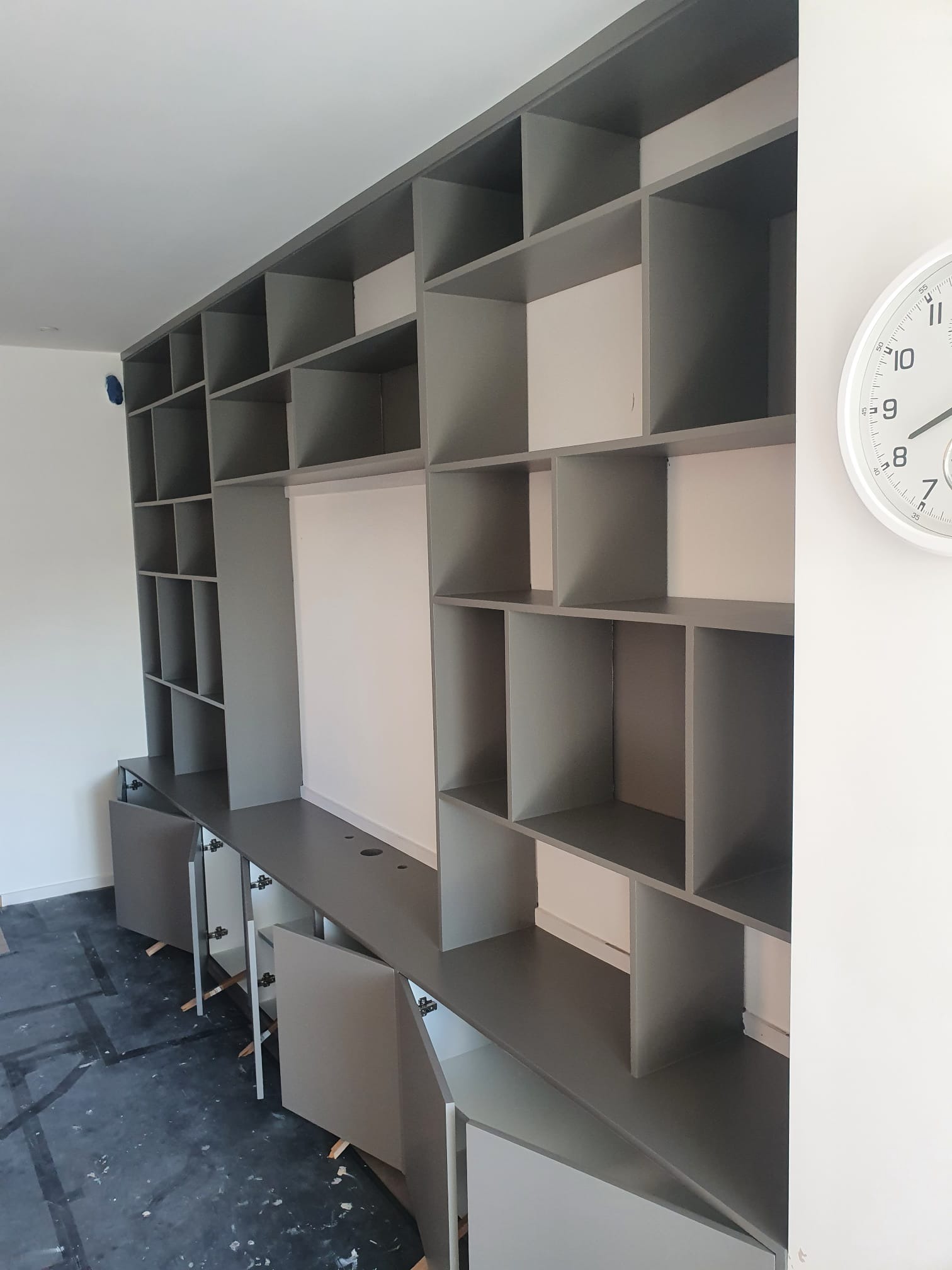 Szaro-biała zabudowa meblowa z otwartymi i zamkniętymi szafkami, widoczne zawiasy i podpórki montażowe, na podłodze folia ochronna, biały zegar na ścianie.