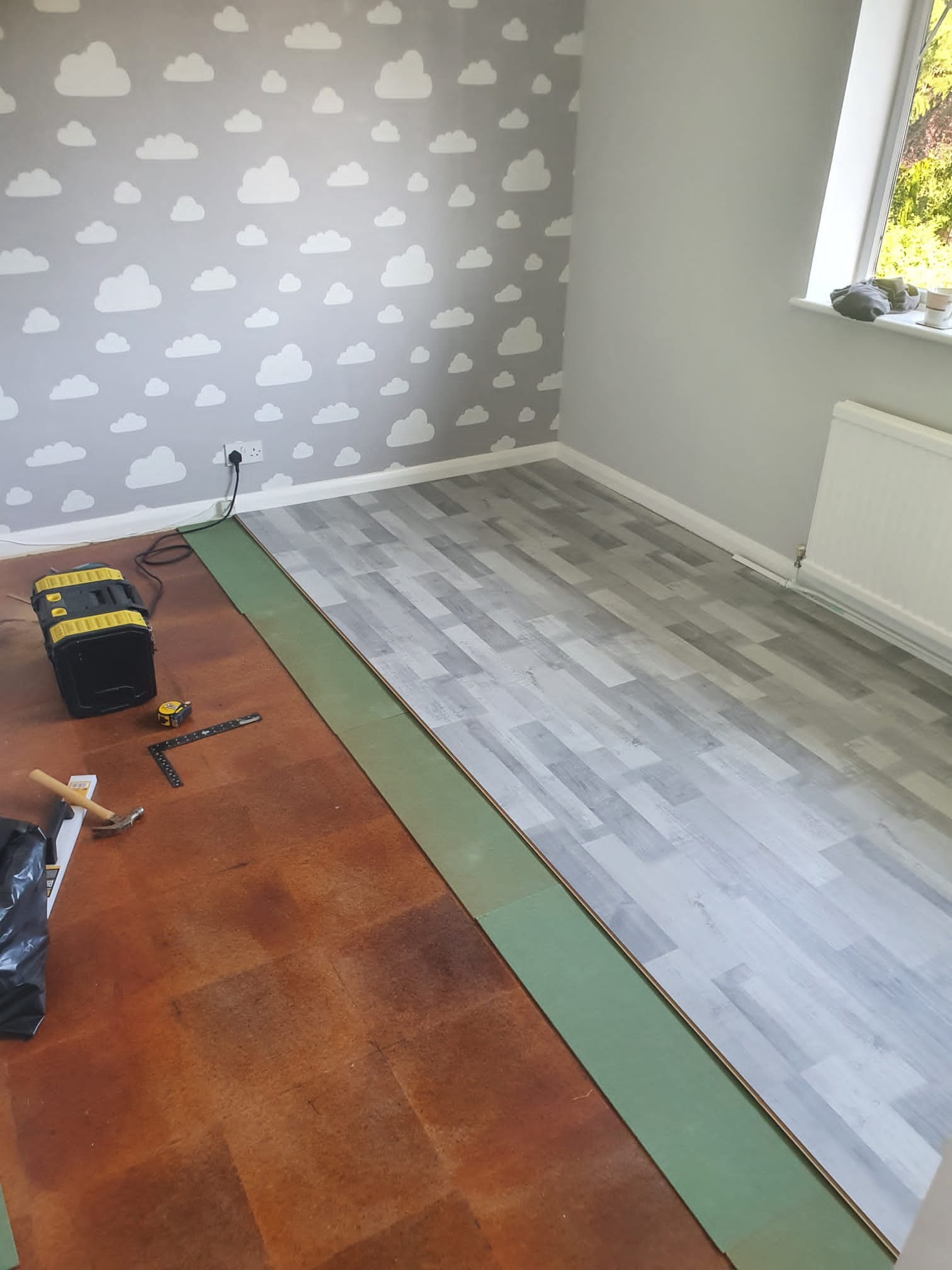 Fragment pokoju z tapetą w chmurki podczas układania paneli podłogowych; widoczna część podłogi z nowymi panelami, narzędzia pracy oraz podkład pod panele.