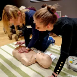 Dwie kobiety ćwiczą resuscytację krążeniowo-oddechową na manekinie na tle ekranu projekcyjnego podczas kursu pierwszej pomocy. Jedna kobieta w ciemnej bluzce z guzikami na rękawach, druga...