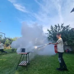 Chłopiec w jasnej koszulce i ciemnych spodniach z uśmiechem gasi pożar grilla za pomocą czerwonej gaśnicy, podczas gdy drugi chłopiec biegnie w oddali, na tle zielonego trawnika i błękitnego nieba.