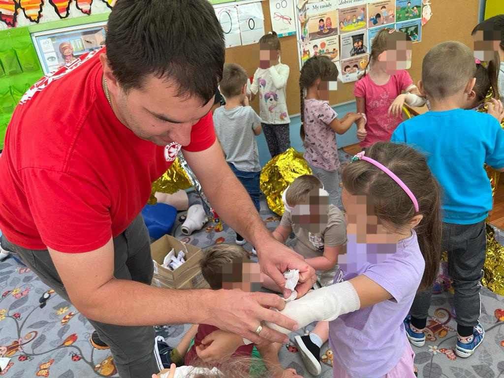 Instruktor w czerwonej koszulce opatruje rękę dziecka bandażem na tle grupy dzieci w sali przedszkolnej.