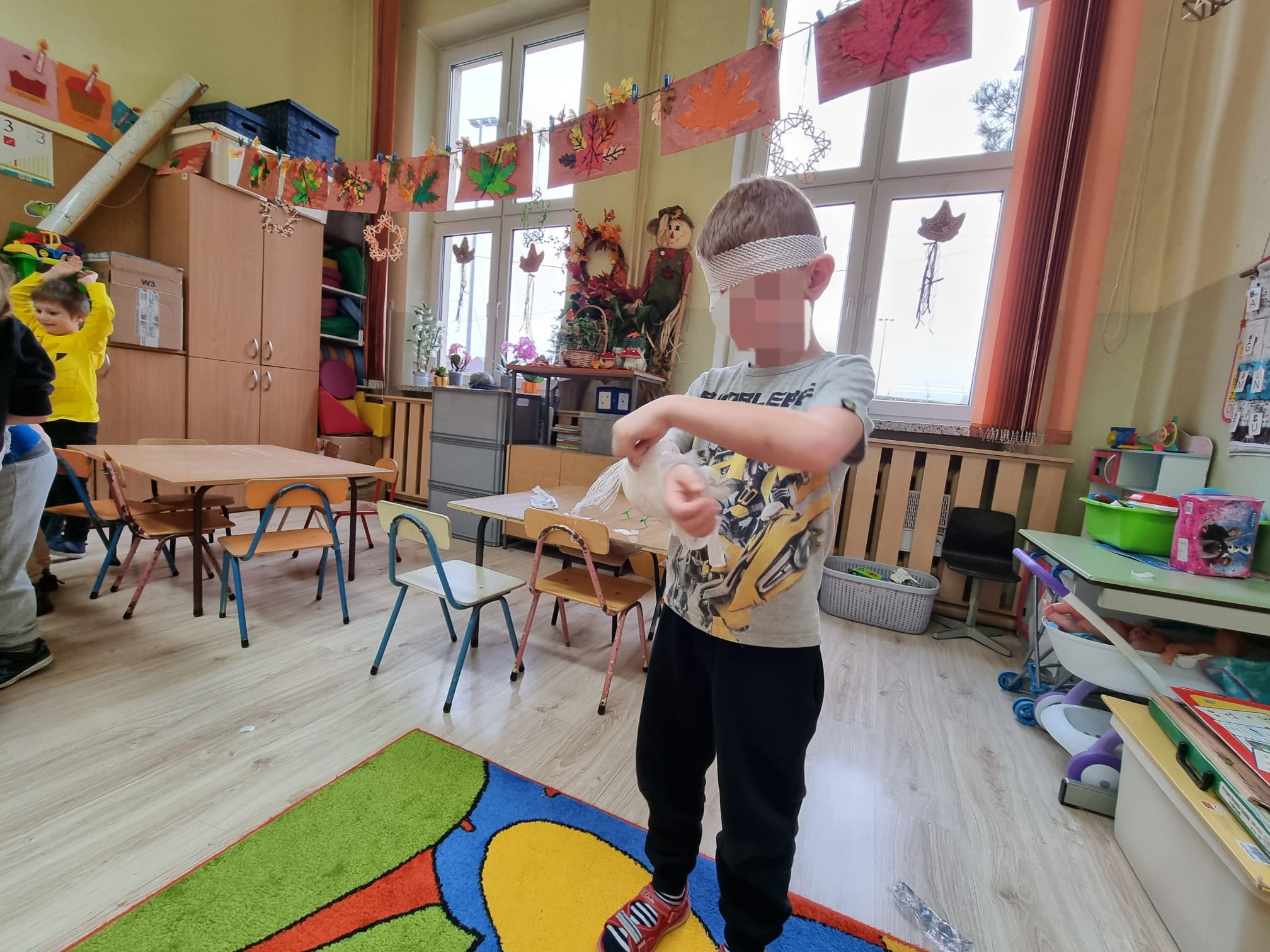 Chłopiec z zasłoniętymi oczami, w przedszkolnej sali, uczy się udzielania pierwszej pomocy, trzymając w rękach przezroczysty woreczek.