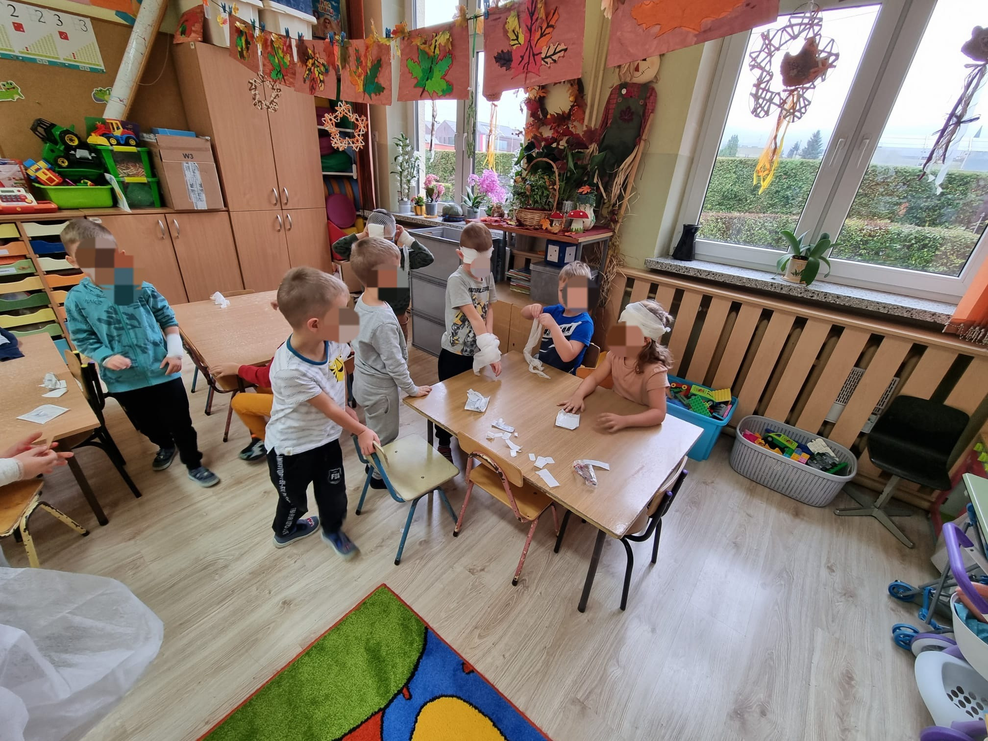 Dzieci w przedszkolu ćwiczą bandażowanie, niektóre z zawiązanymi oczami, siedząc przy drewnianym stole z porozrzucanymi kawałkami bandaży.