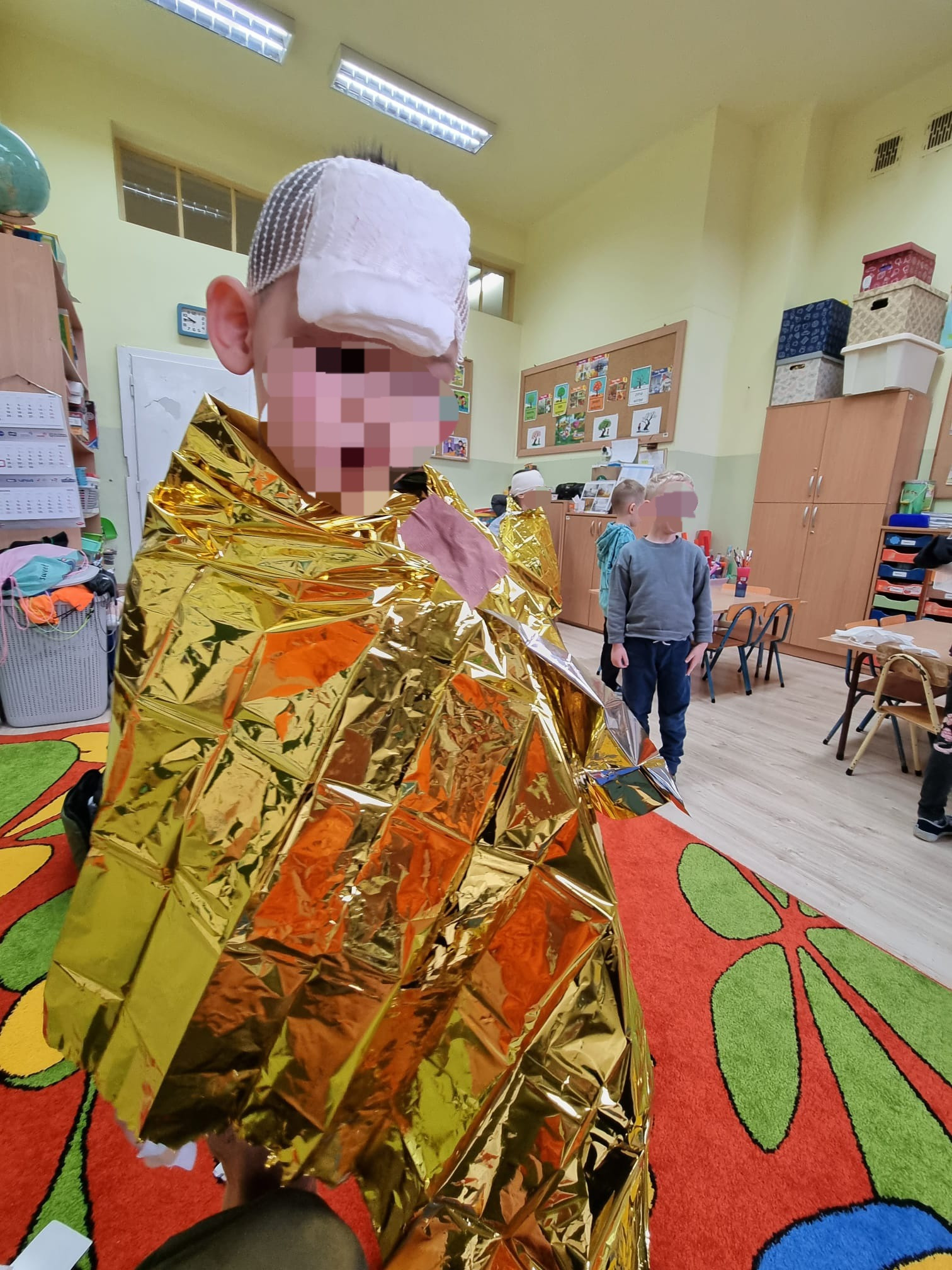 Chłopiec z opatrunkiem na głowie, owinięty złotą folią termiczną w sali przedszkolnej, symulacja udzielania pierwszej pomocy.