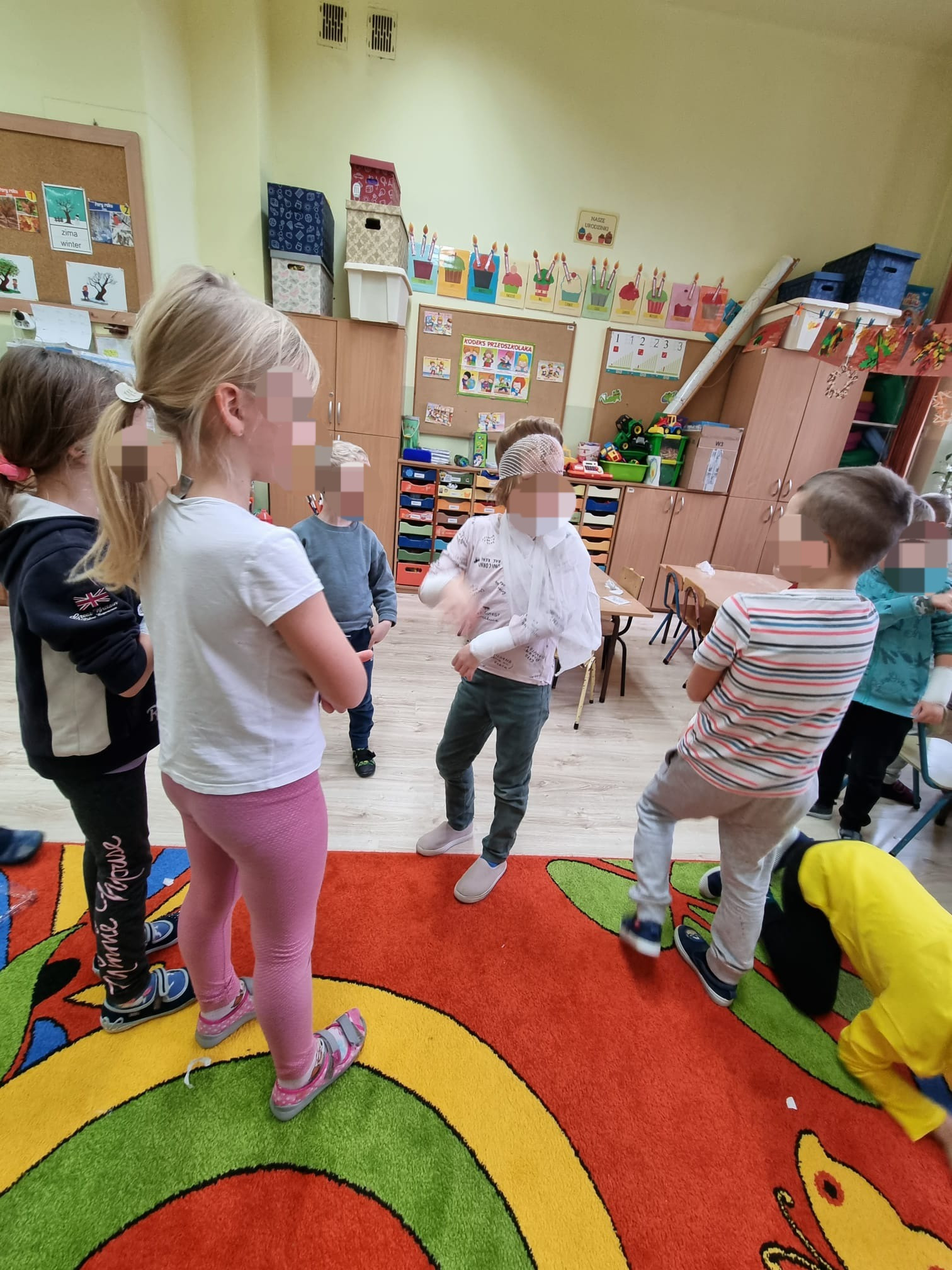 Dzieci w sali przedszkolnej uczestniczą w symulacji udzielania pierwszej pomocy, jedno dziecko ma założony bandaż na głowie i ręce.