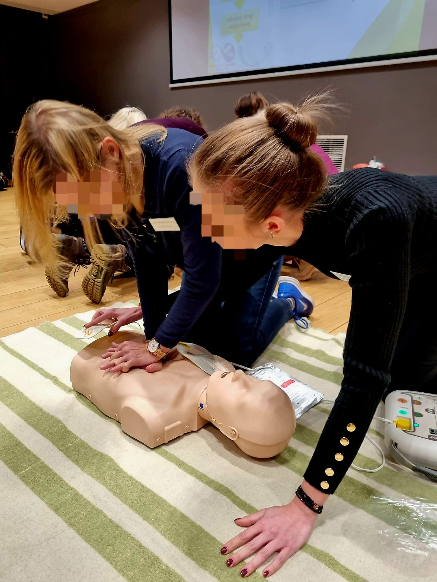 Dwie kobiety ćwiczą resuscytację krążeniowo-oddechową na manekinie na tle ekranu projekcyjnego podczas kursu pierwszej pomocy. Jedna kobieta w ciemnej bluzce z guzikami na rękawach, druga...